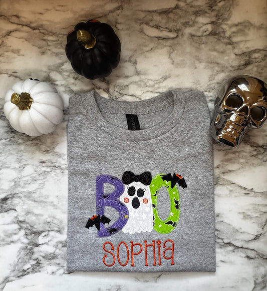 Gildan Youth T-Shirt with a Boo Embroidered Design where each letter has Halloween themed fabric and a cute ghost replaces one of the "O"s in the word.  Shirt can be personalized with child's name for extra cuteness.