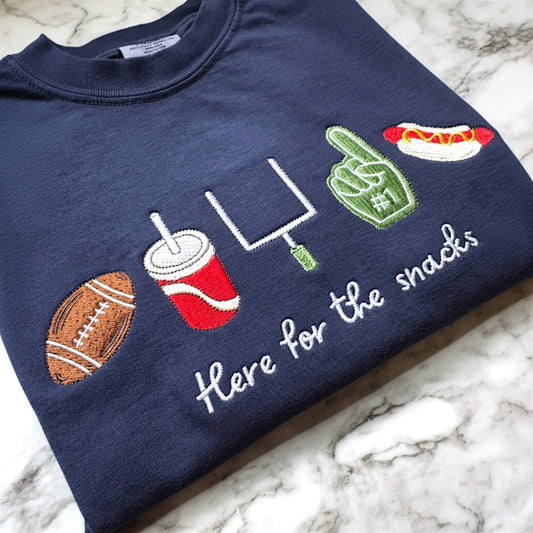 Navy Comfort Colors football game day t-shirt folded on a tabletop featuring embroidered “Here for the Snacks” football and snack icons.
