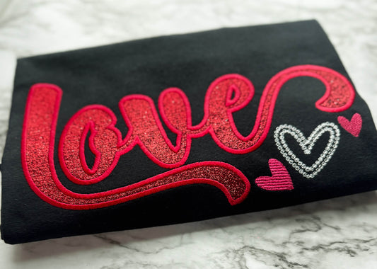 Gildan Black T-Shirt with the word "Love" in a script font embroidered in two different color options with glyphs added to some of the letters.  This shirt uses red glitter HTV with matching red stitching around the border of the word.  It also has small hearts in white and pink as accents to the side of the word.