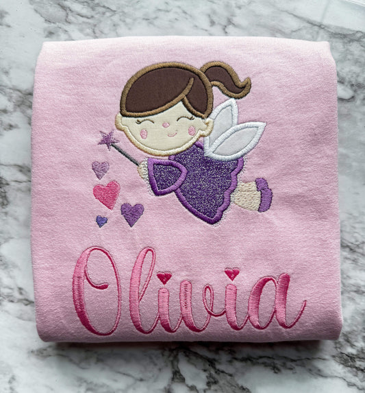 Gildan Light Pink TShirt with the cutest fairy embroidered design.  The fairy is dressed in a purple glittery dress holding a magic wand with hearts in the colors pink and purple cascading down.  You can personalize the shirt by adding your child's name in the color pink.  Font used will be dependent on the length of the name.