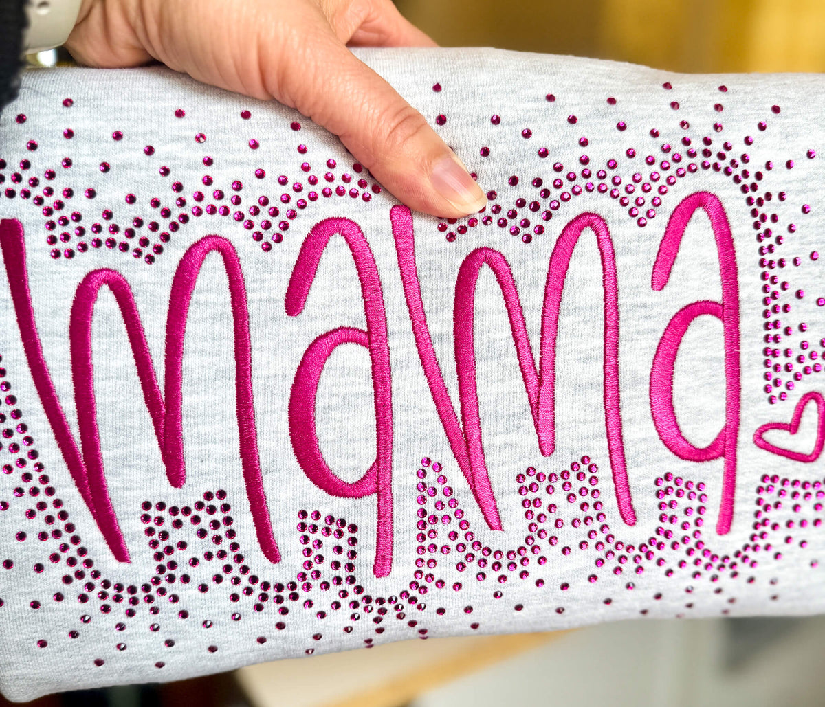 Gildan Softstyle Sweatshirt with the word "MAMA" embroidered in a large font.  The sweatshirt colors offered are:  1) Ash Gray with hot pink stitching, 2) Black with red stitching, 3) Lt Pink with pink stitching and 4) Navy with medium blue stitching.  Each shirt comes with the option of adding a rhinestone splatter around the embroidery in a complimentary color.