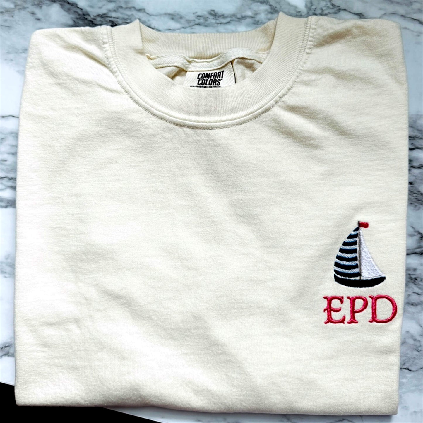 Ivory Comfort Colors t-shirt featuring a left chest embroidered sailboat with your embroidered initials. Part of our Coastal Collection, this customizable embroidered tee can be personalized with your favorite beach town, island, coastal destination, or initials (only the sailboat has the option of initials) and is available in five coastal-inspired colors.