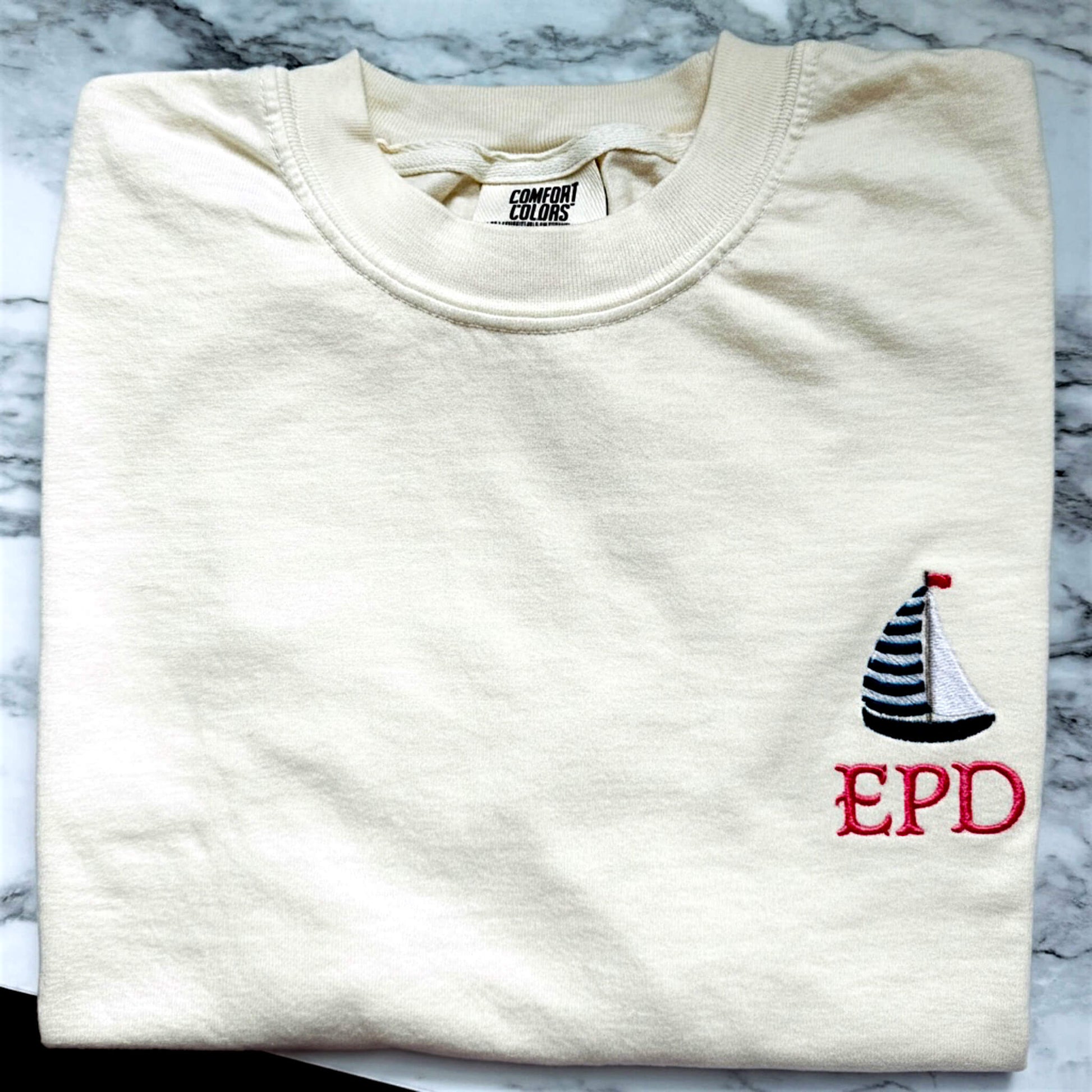 Ivory Comfort Colors t-shirt featuring a left chest embroidered sailboat with your embroidered initials. Part of our Coastal Collection, this customizable embroidered tee can be personalized with your favorite beach town, island, coastal destination, or initials (only the sailboat has the option of initials) and is available in five coastal-inspired colors.