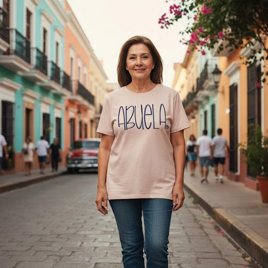 Model wearing a Gildan T-Shirt in the color Dusty Rose with the word "Abuela" embroidered in a large handwritten font.  The color of the thread compliments the shirt color.