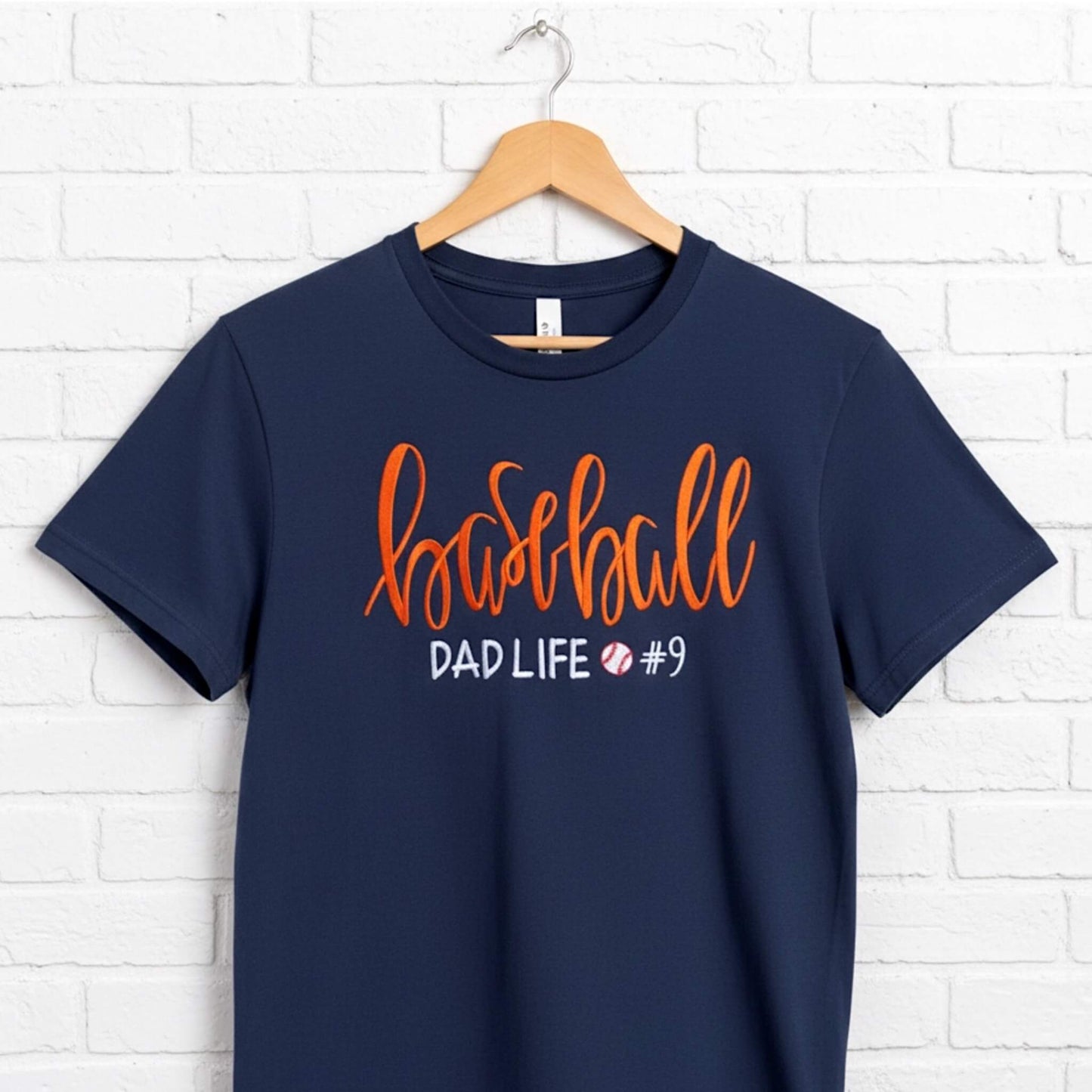 Gildan navy blue T-shirt embroidered with the following:  the word "Baseball" in a large script handwritten font, with the tag line Dad Life and a cute heart.  You have the option to pick your shirt color, add a jersey number and pick a thread color for the word baseball to coordinate with your team's colors.