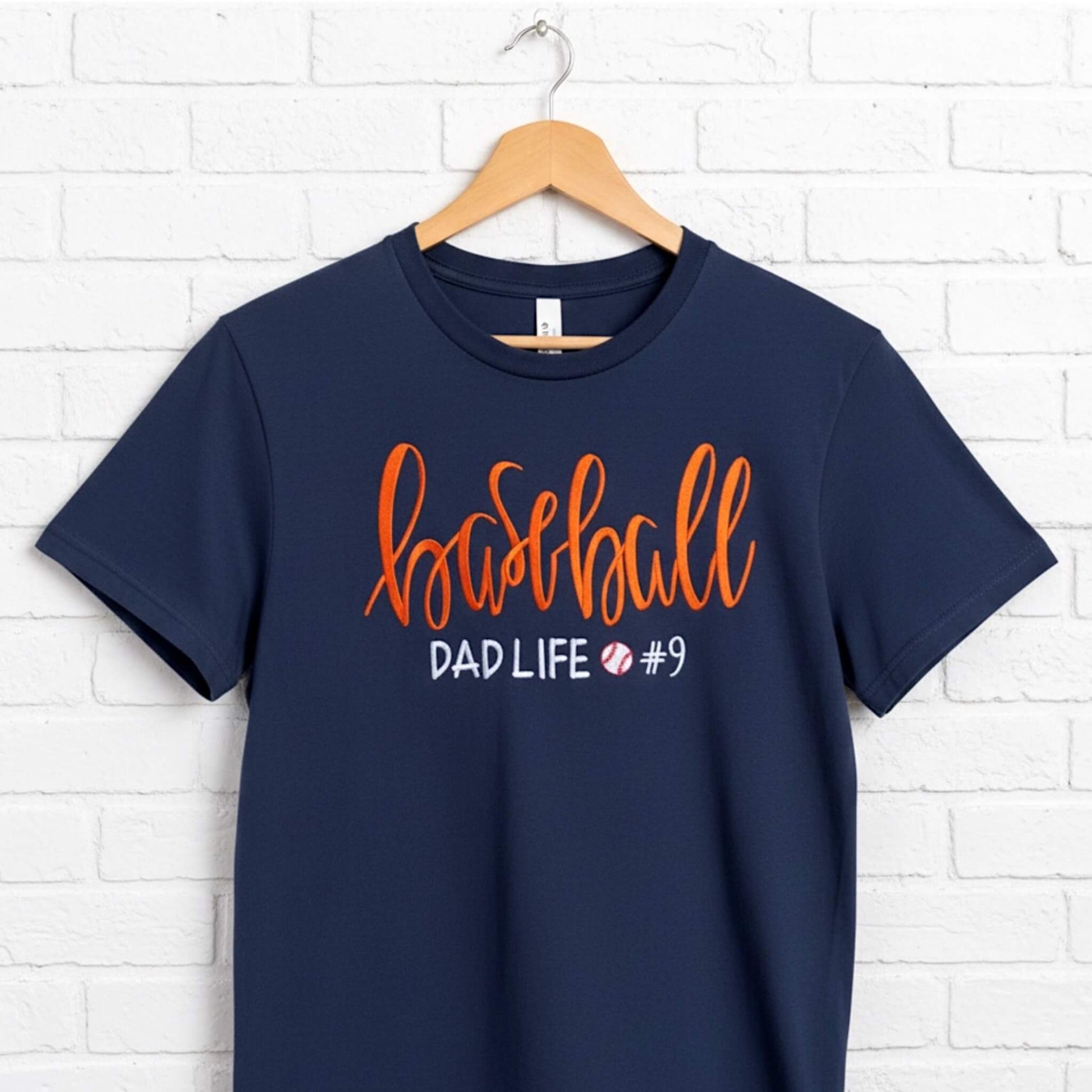 Gildan navy blue T-shirt embroidered with the following:  the word "Baseball" in a large script handwritten font, with the tag line Dad Life and a cute heart.  You have the option to pick your shirt color, add a jersey number and pick a thread color for the word baseball to coordinate with your team's colors.