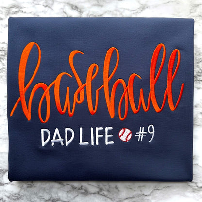 Gildan navy blue T-shirt embroidered with the following:  the word "Baseball" in a large script handwritten font, with the tag line Dad Life and a baseball.  You have the option to pick your shirt color, add a jersey number and pick a thread color for the word baseball to coordinate with your team's colors.