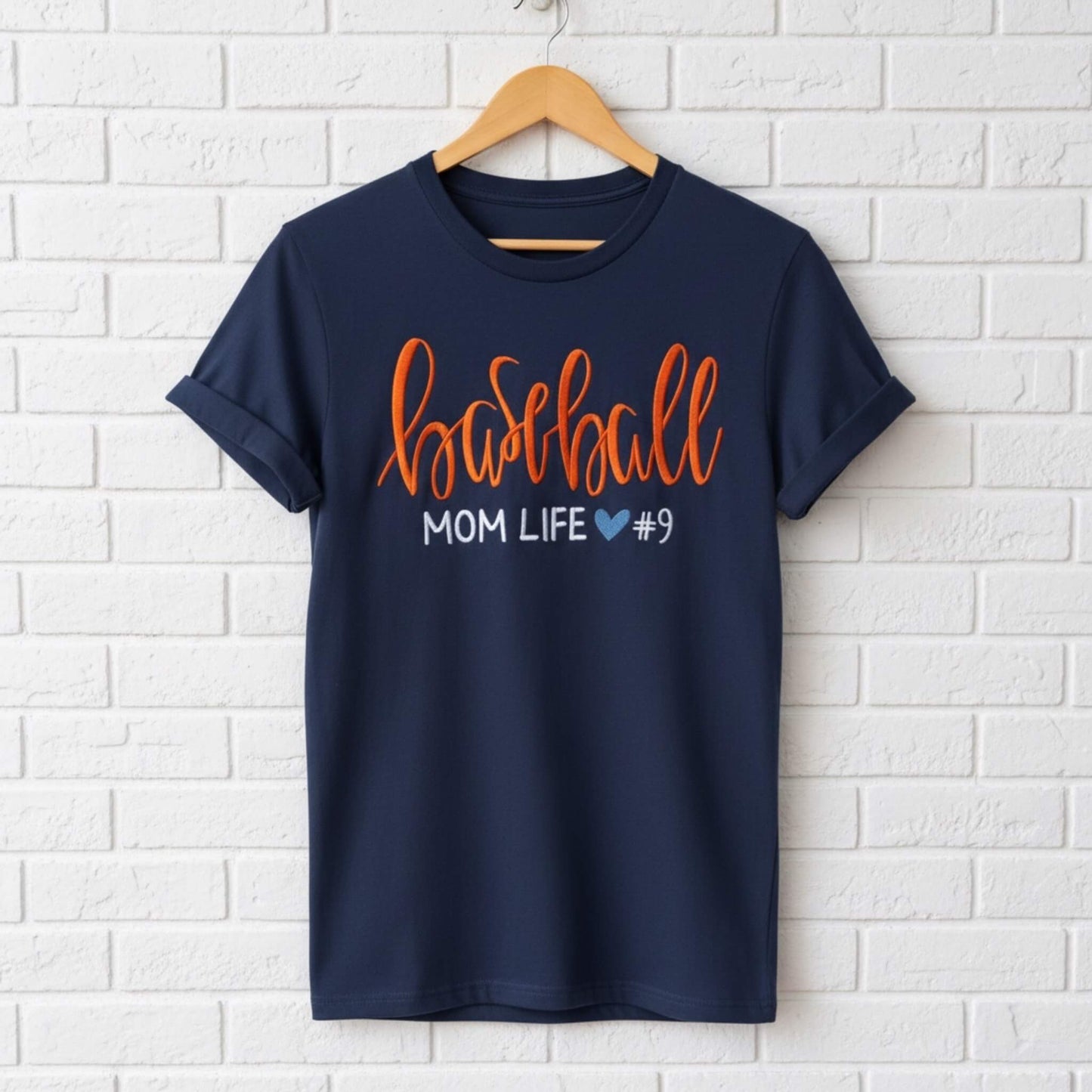 Gildan navy blue T-shirt embroidered with the following:  the word "Baseball" in a large script handwritten font, with the tag line Mom Life and a cute heart.  You have the option to pick your shirt color, add a jersey number and pick a thread color for the word baseball to coordinate with your team's colors.