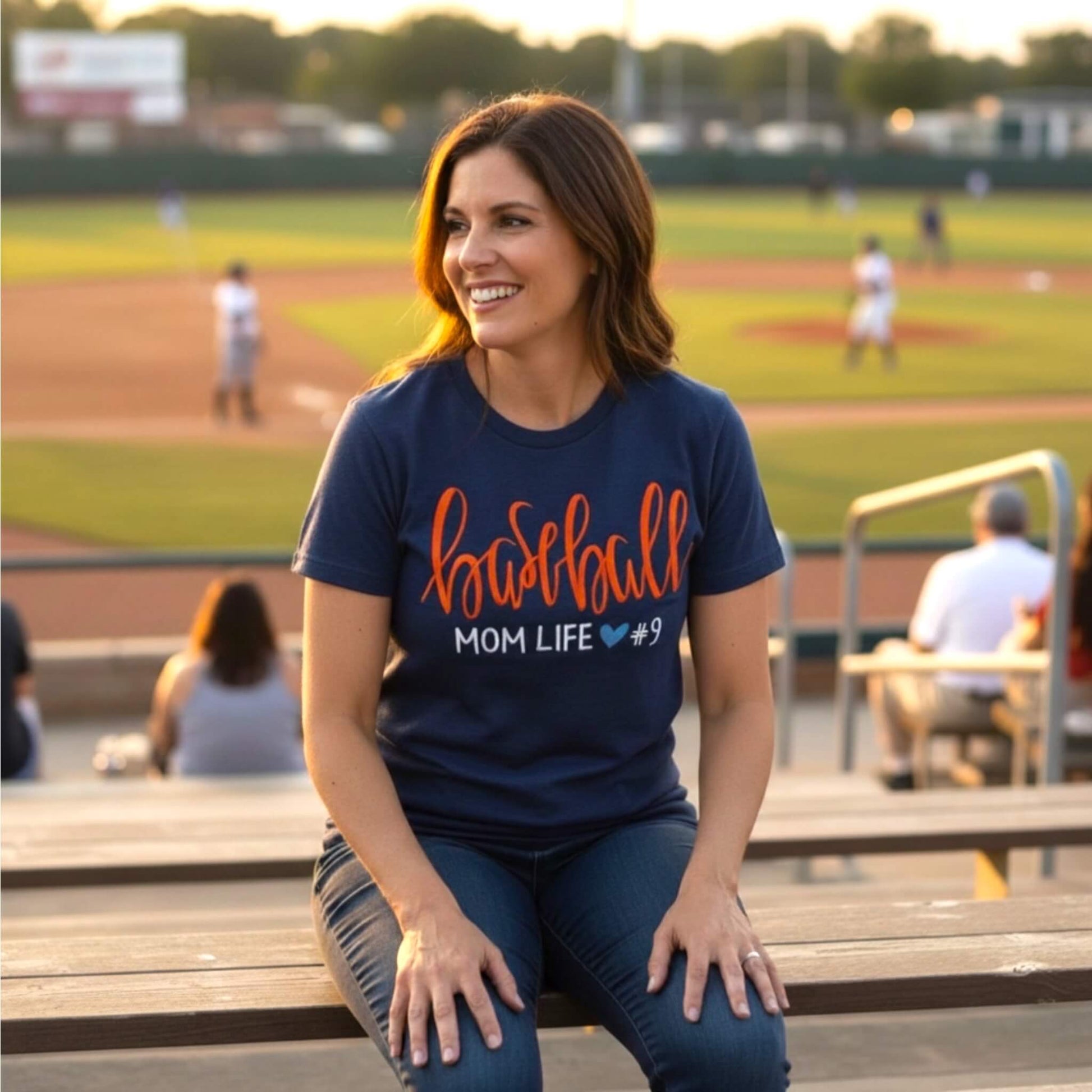 Model wearing a Gildan navy blue T-shirt embroidered with the word "Baseball" in a large script handwritten font, with the tag line Mom Life and her child's jersey number.  