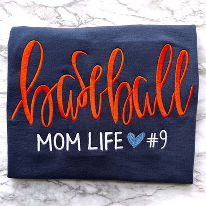 Gildan navy blue T-shirt embroidered with the following:  the word "Baseball" in a large script handwritten font, with the tag line Mom Life and a cute heart.  You have the option to pick your shirt color, add a jersey number and pick a thread color for the word baseball to coordinate with your team's colors.