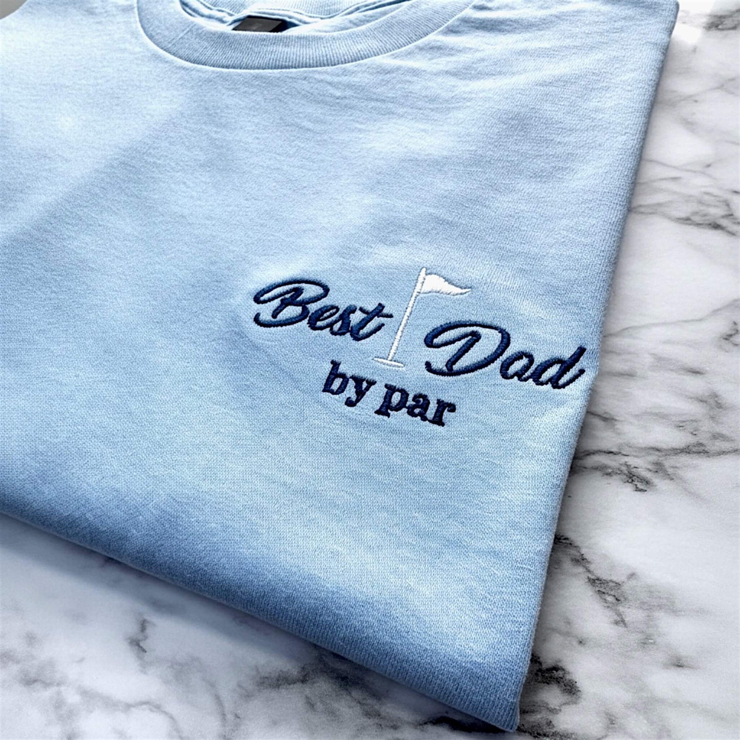 Close up image of a Gildan Light Blue T-Shirt with a left chest embroidered design that says: "Best Dad by Par" with a golf flag.