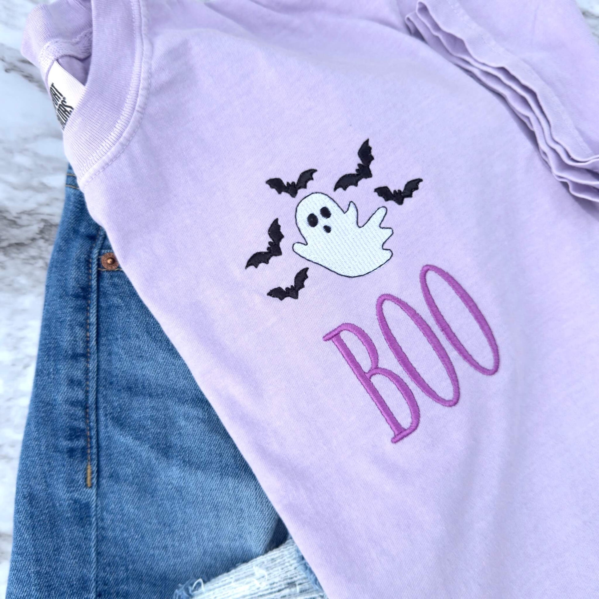 Close-up of embroidered ghost, bats and BOO Halloween design showing detailed stitching.  Design is on a Comfort Colors T-shirt in the color Orchid.  Styled with a pair of jean shorts, which are not included.