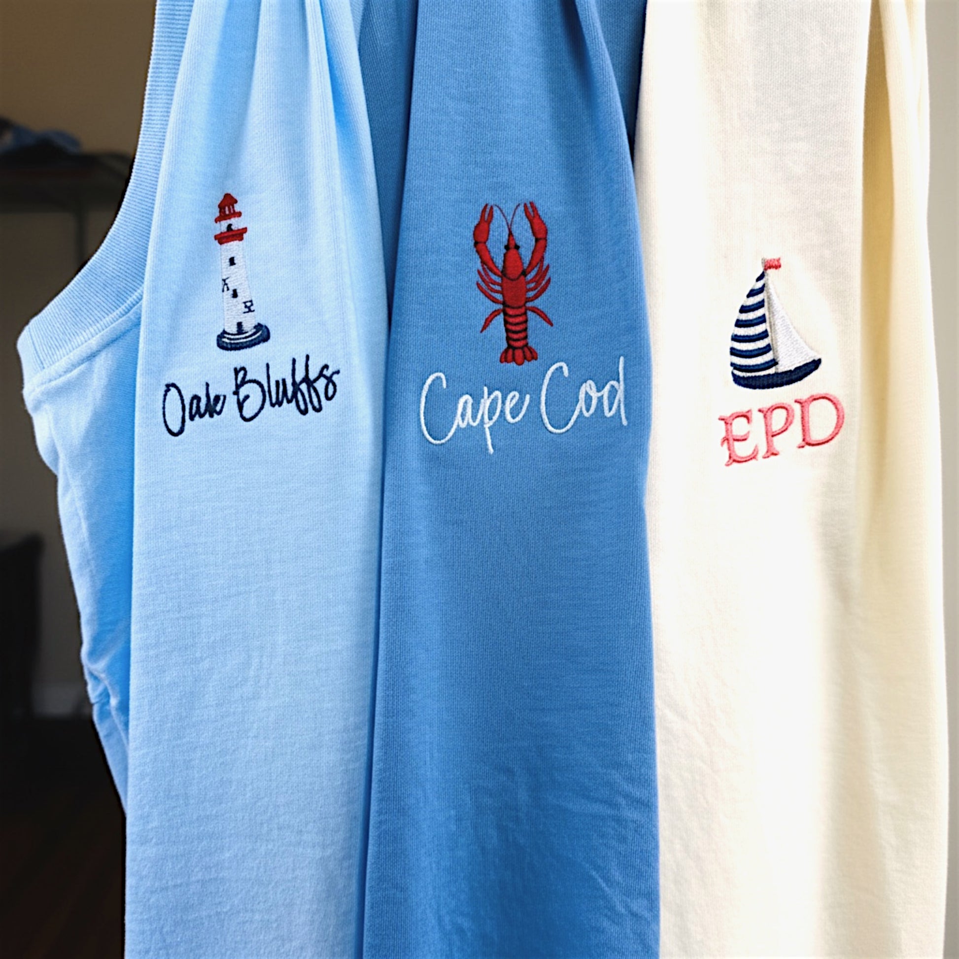 Three embroidered Comfort Colors t-shirts hanging together from our Coastal Collection, featuring personalized lobster, sailboat, and lighthouse designs inspired by coastal destinations and nautical living.