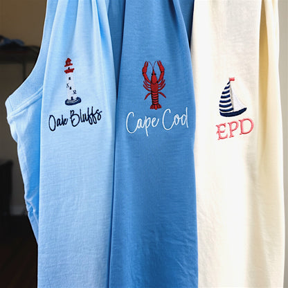 Three embroidered Comfort Colors t-shirts hanging together from our Coastal Collection, featuring personalized lobster, sailboat, and lighthouse designs inspired by coastal destinations and nautical living.
