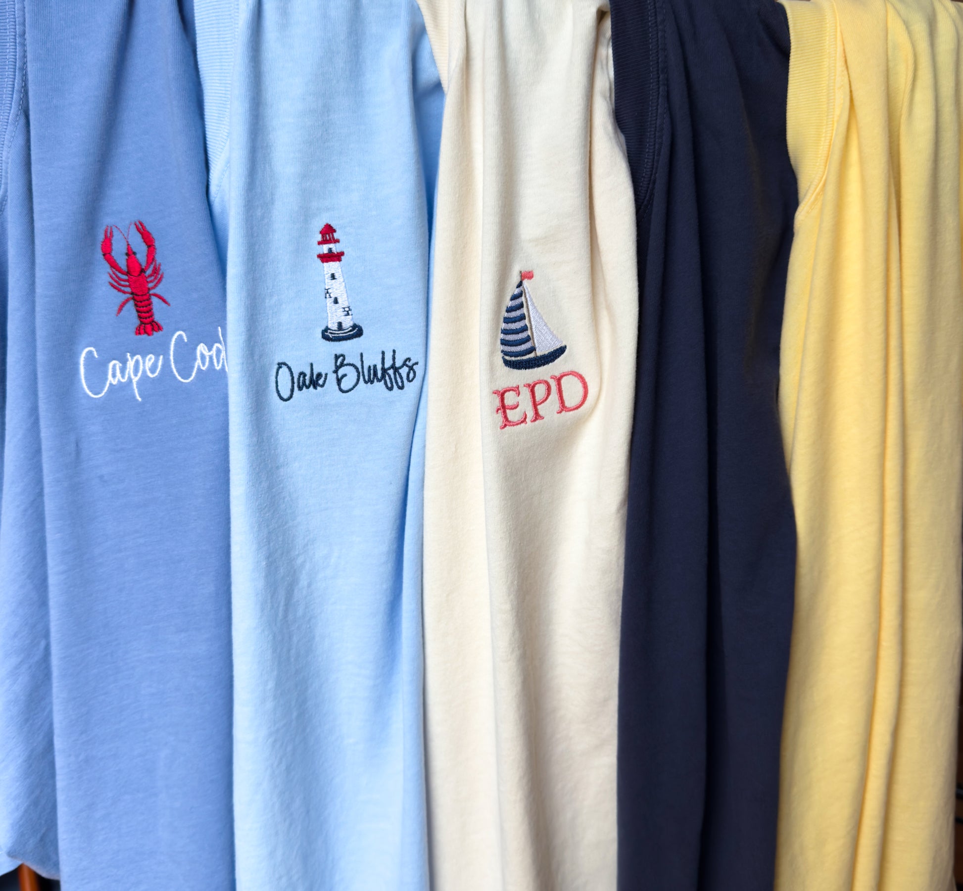 Three embroidered Comfort Colors t-shirts hanging together from our Coastal Collection, featuring personalized lobster, sailboat, and lighthouse designs inspired by coastal destinations and nautical living.  The two other available colors for the collection are shown.