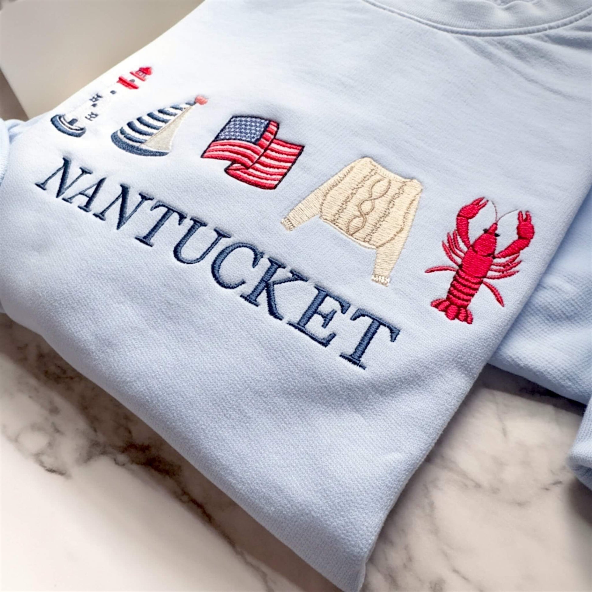 Close-up of premium embroidery on a personalized Comfort Colors lightweight sweatshirt in the color Hydrangea featuring detailed coastal icons and a custom destination location.