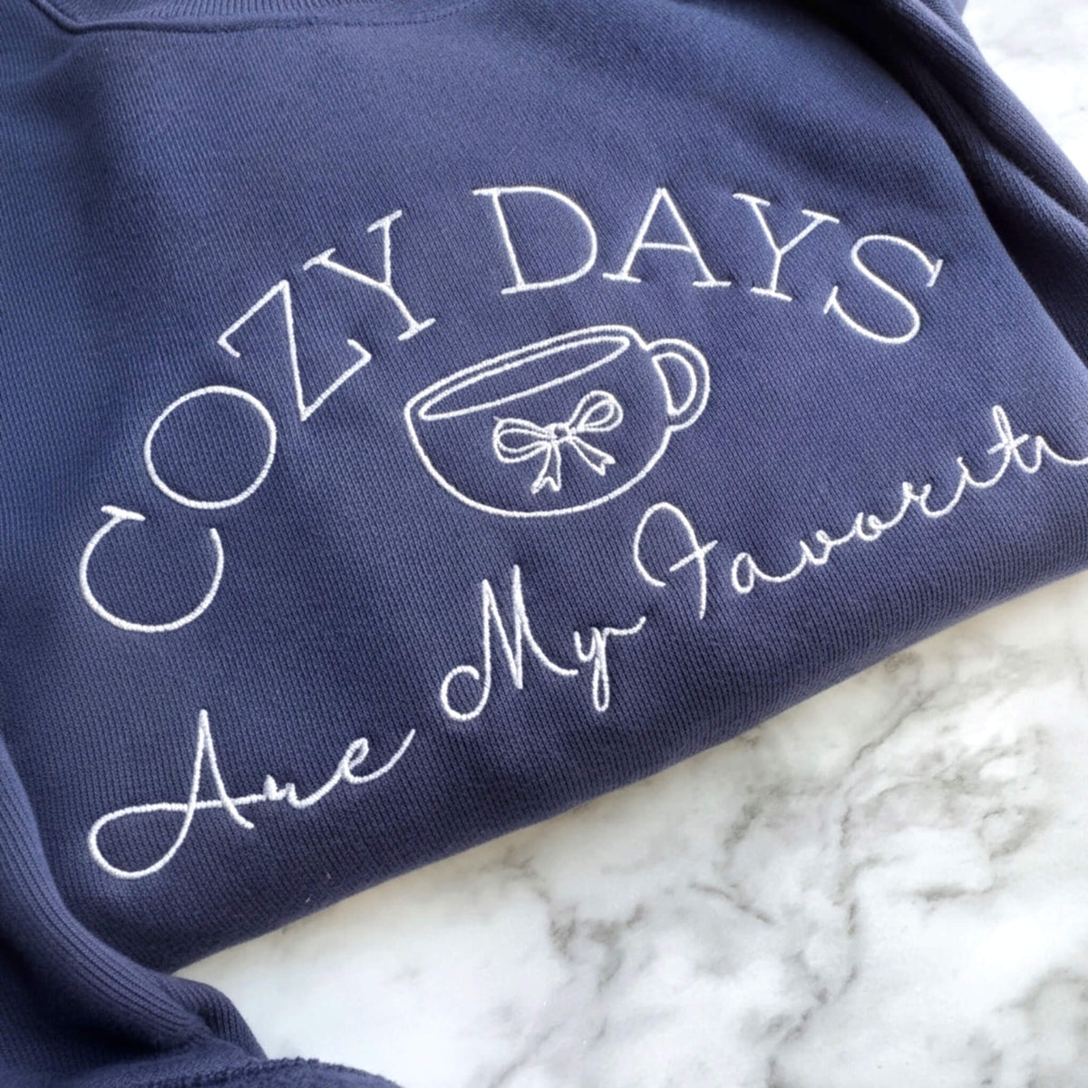 Close up view of a navy Bella + Canvas crewneck sweatshirt with white Cozy Days Are My Favorite embroidered design featuring a coffee cup and bow.