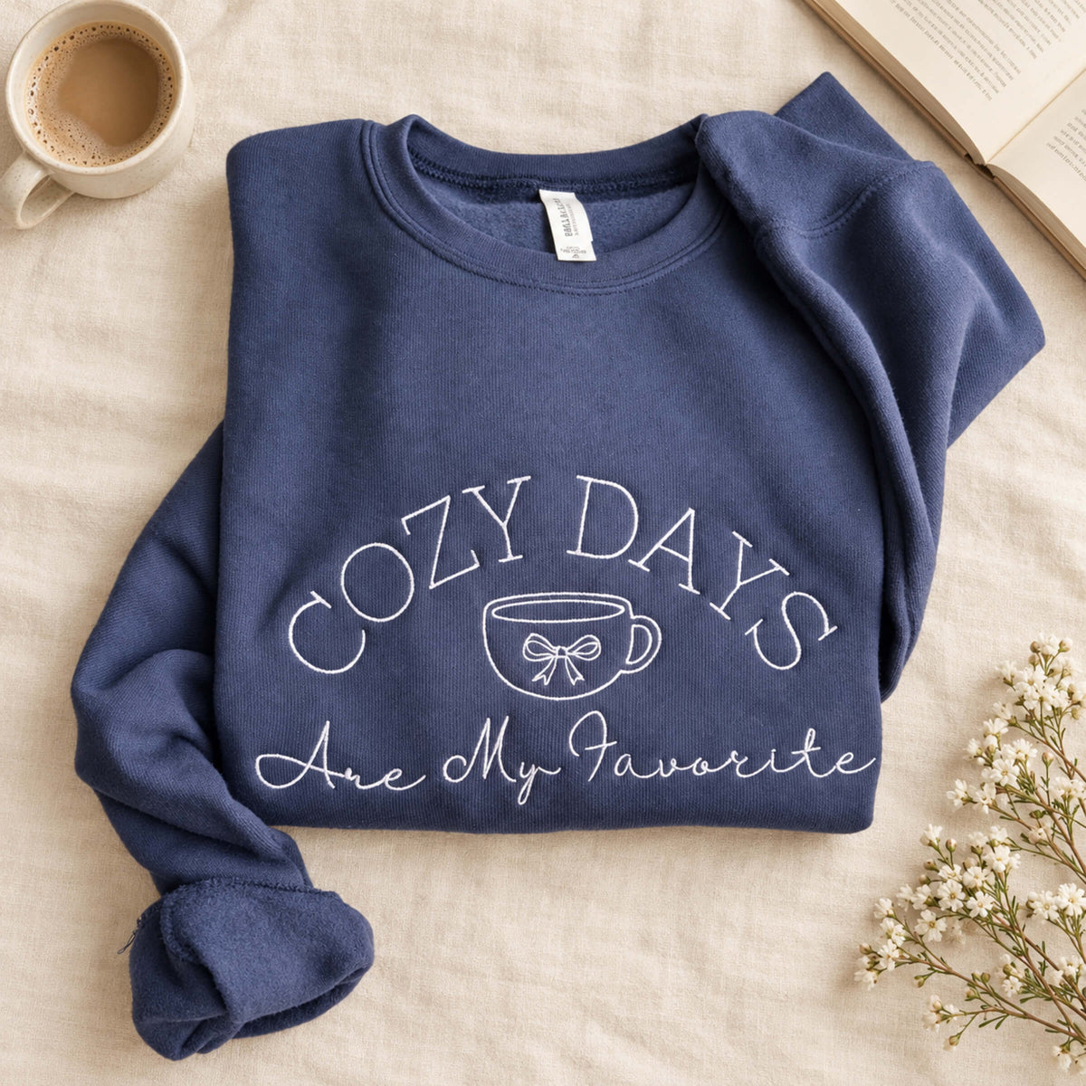 Lifestyle flat lay of a navy Bella + Canvas crewneck sweatshirt with white Cozy Days Are My Favorite embroidered design featuring a coffee cup and bow.