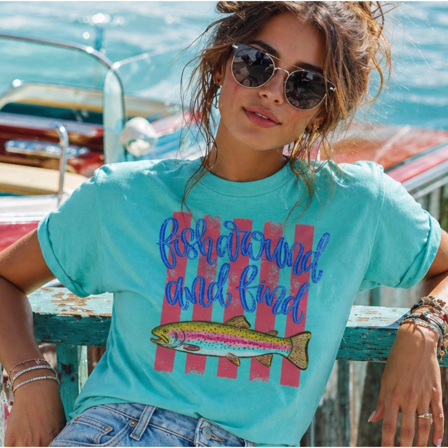 Model wearing a Comfort Colors tee with a hand-drawn trout fish graphic with hot pink horizontal lines and script text saying: "Fish Around and Find Trout" in the color Lagoon.