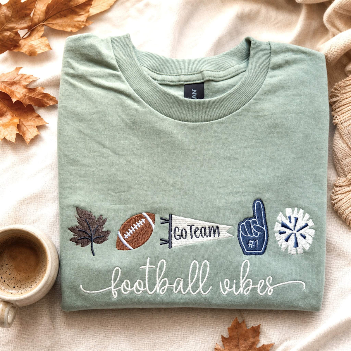 Gildan Softstyle Sage green embroidered football vibes T-shirt styled in a fall game day flat lay with football-themed accessories.