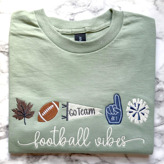 Sage green Gildan Softstyle T-shirt folded to show an embroidered football themed design with fall and game day icons and the word "football vibes" in a script font.