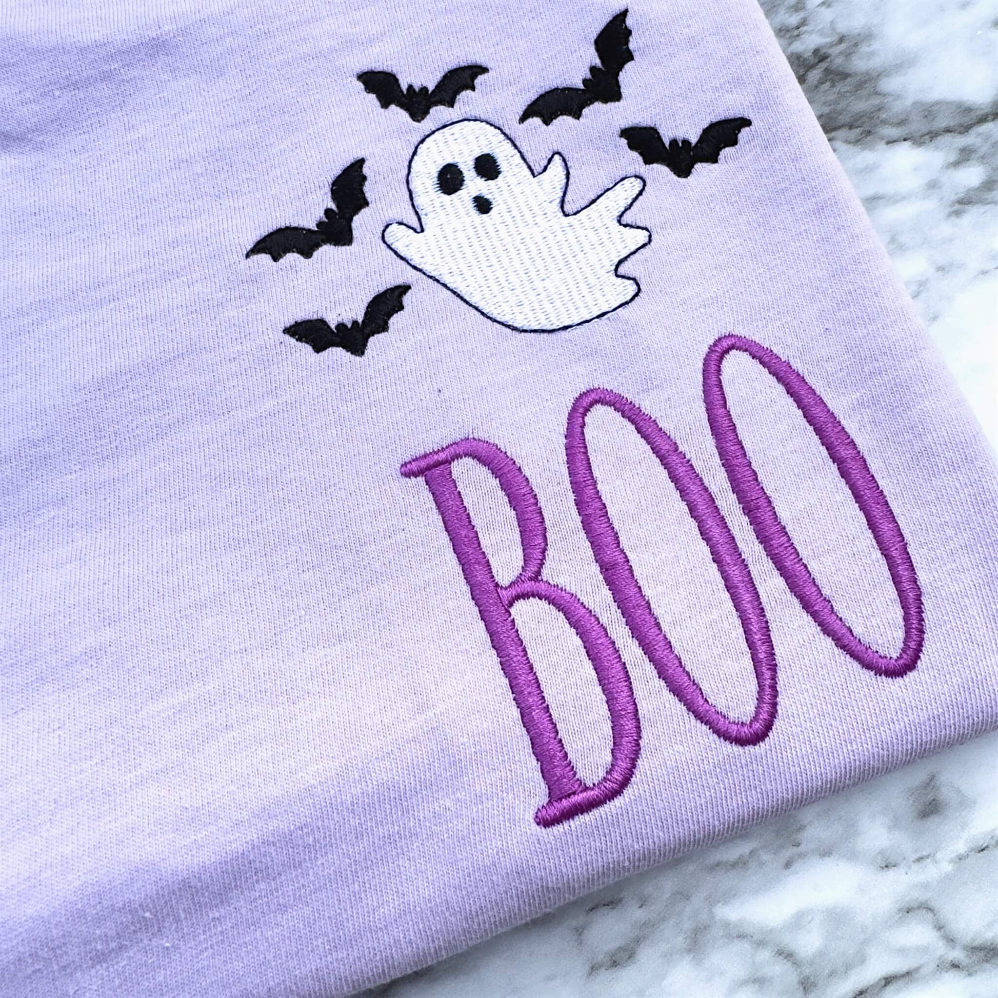 Close-up of embroidered ghost, bats and BOO Halloween design showing detailed stitching.  Design is on a Comfort Colors T-shirt in the color Orchid.