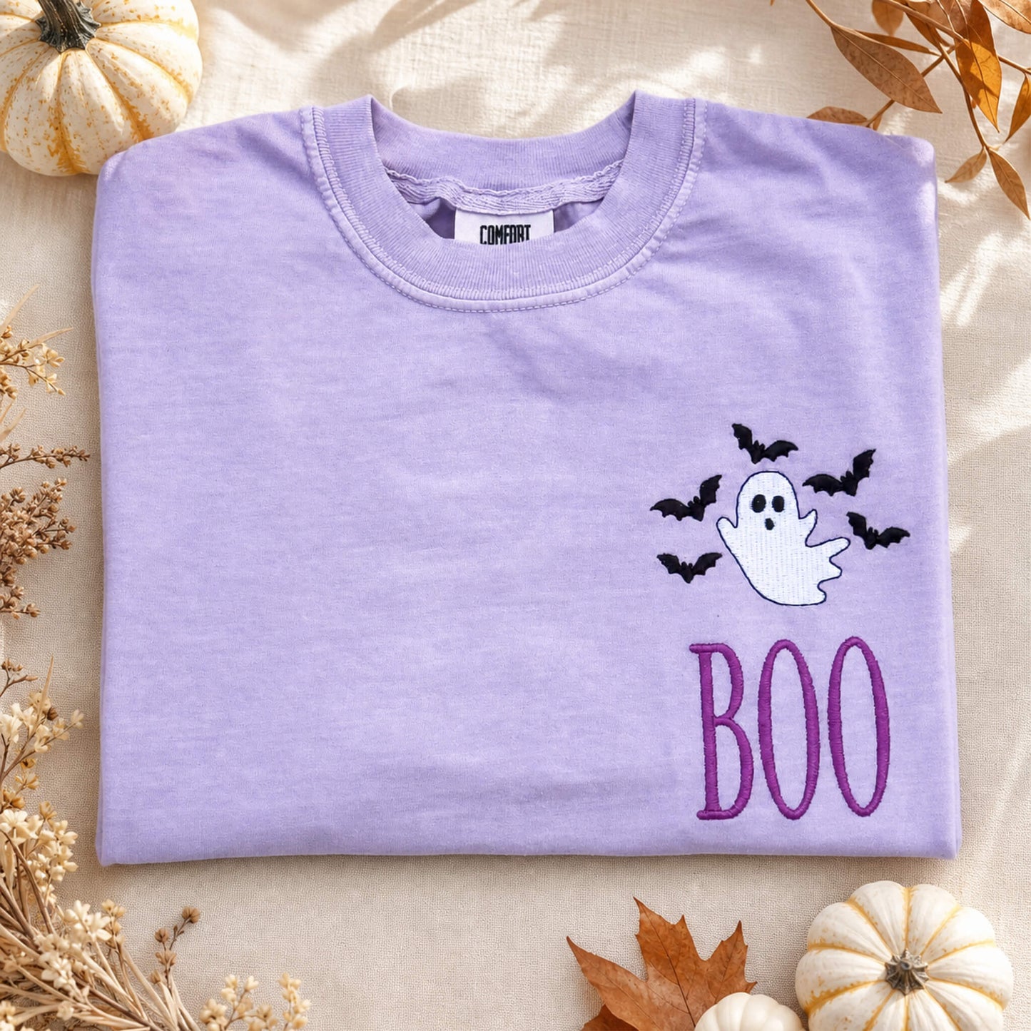 Ghost and bats BOO embroidered Halloween design on a Comfort Colors tee in the color Orchid styled with fall decorations.