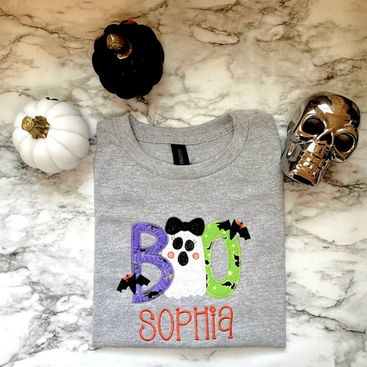 Gildan Youth T-Shirt in the color Sports Gray with a Boo Embroidered Design where each letter has Halloween themed fabric and a cute ghost replaces one of the "O"s in the word.  Shirt can be personalized with child's name for extra cuteness.