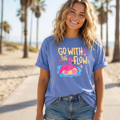 An AI model wearing a Comfort Colors T-Shirt in the color Mystic Blue with a cute design that shows a hot pink flamingo hanging out on a pool floaty with the words "Go with the Flow" across the top.  Perfect for summer.
