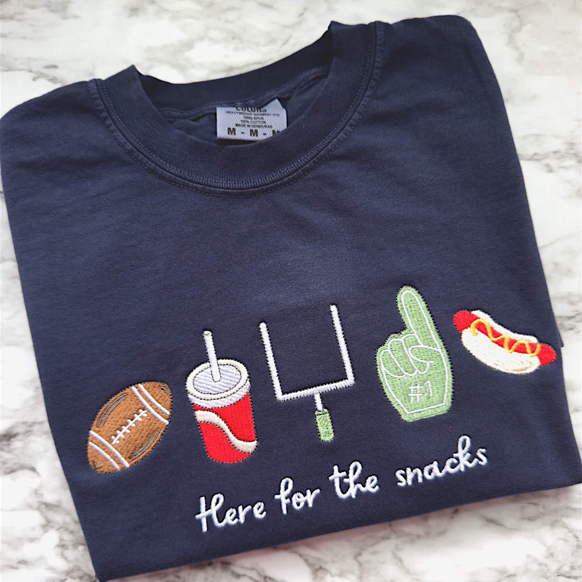 Navy Comfort Colors football game day t-shirt folded on a tabletop featuring embroidered “Here for the Snacks” football and snack icons.