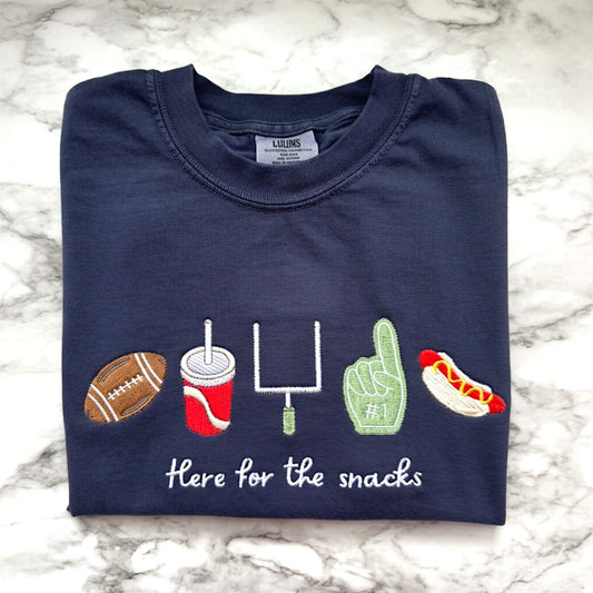 Navy Comfort Colors football game day t-shirt folded on a tabletop featuring embroidered “Here for the Snacks” football and snack icons.