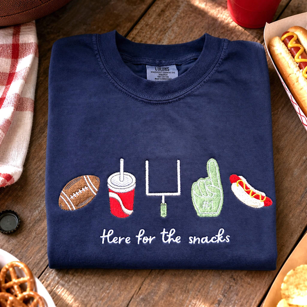 Navy Comfort Colors “Here for the Snacks” football t-shirt folded in a casual game day flat lay with a football and snacks.