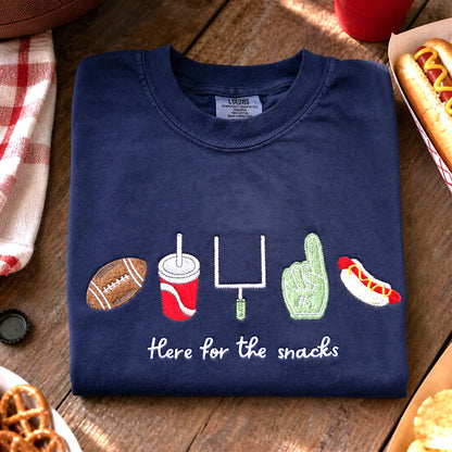 Navy Comfort Colors “Here for the Snacks” football t-shirt folded in a casual game day flat lay with a football and snacks.