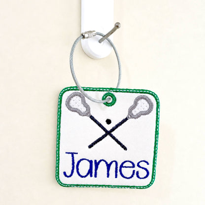 A personalized embroidered Lacrosse themed bag tag featuring crossed lacrosse sticks, bordered by green stitching and hanging from a silver wire loop on a white wall hook.  Personalize with your child's name in the thread color of your choice.