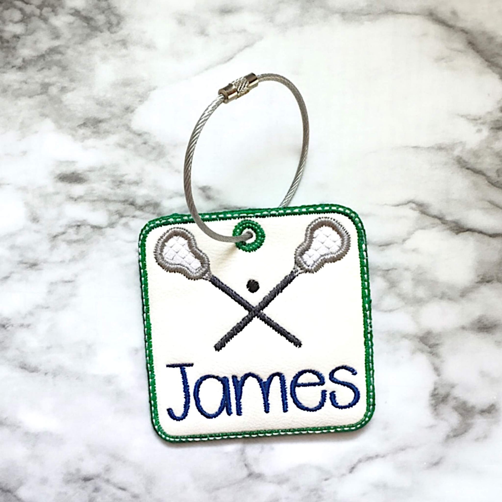 A square embroidered bag tag with a stainless steel twist closure wire ring, featuring crossed lacrosse sticks, a green stitched border, and the name 'James' in blue on white marine vinyl, placed on a gray and white marble surface.