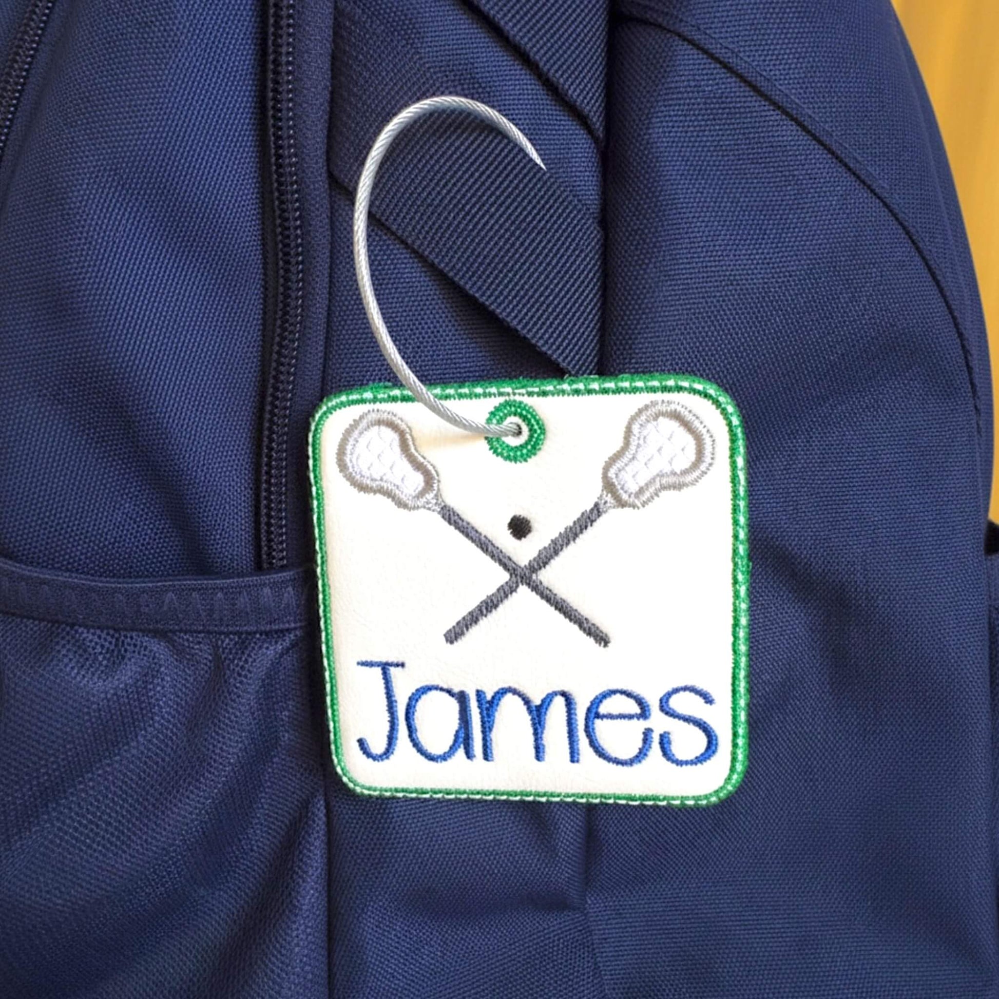 A close-up of a navy blue backpack with a personalized embroidered lacrosse bag tag. The bag tag is white with a green border, featuring crossed lacrosse sticks and the name 'James' in blue text, hanging from a stainless steel twist closure ring.