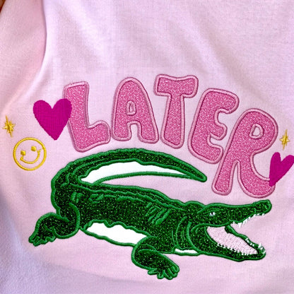 Close up look at a Gildan Light Pink T-Shirt with an applique embroidered design with the word "Later" and an alligator underneath it as a throw back to the old school saying "See ya later alligator".  The design uses HTV glitter in the colors pink and green with cute embroidered accents in hot pink and yellow.