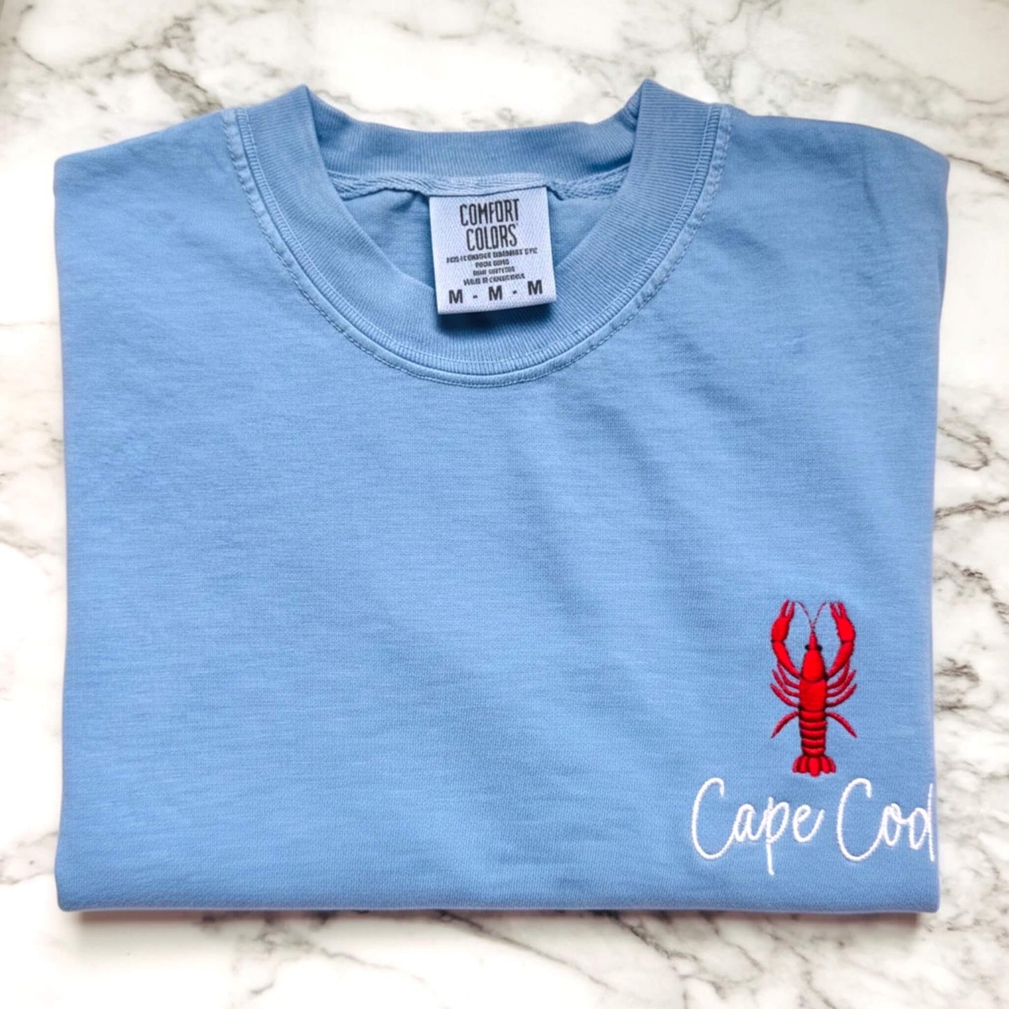 Comfort Colors t-shirt featuring a left chest embroidered lobster with the words Cape Cod.  Part of our Coastal Collection, this customizable embroidered tee can be personalized with your favorite beach town, island, coastal destination, or initials (sailboat design only) and is available in five coastal-inspired colors.