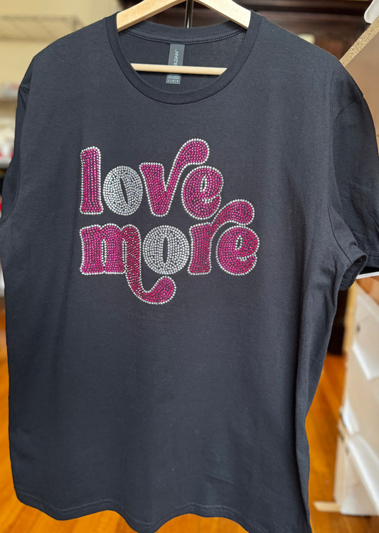 Image is of a black Gildan Softstyle T-Shirt with a rhinestone design that says "Love More" in the rhinestone color magenta while the border uses crystal stones.  The font uses special glyphs on certain letters to make it look unique.  The letter "O" in each word is in all crystal rhinestones instead of magenta.  The design can be purchased on a Bella Canvas T-Shirt or a Gildan Softstyle.