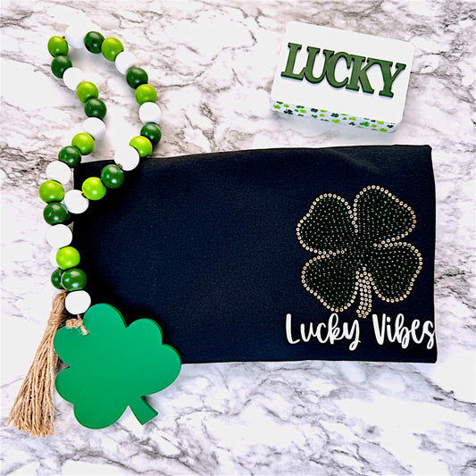 Black T-Shirt with a left chest design.  Design is a St. Patrick's Day Clover in Emerald green and gold rhinestones with the words "Lucky Vibes" in HTV underneath it in a script font.  The design can be put on a Bella Canvas or Gildan Softstyle T-Shirt.