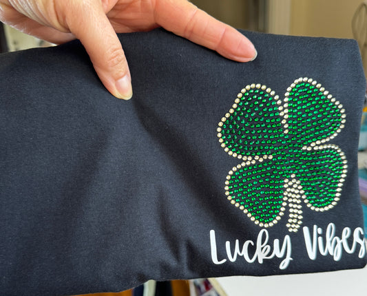 Black T-Shirt with a left chest design.  Design is a St. Patrick's Day Clover in Emerald green and gold rhinestones with the words "Lucky Vibes" in HTV underneath it in a script font.  The design can be put on a Bella Canvas or Gildan Softstyle T-Shirt.