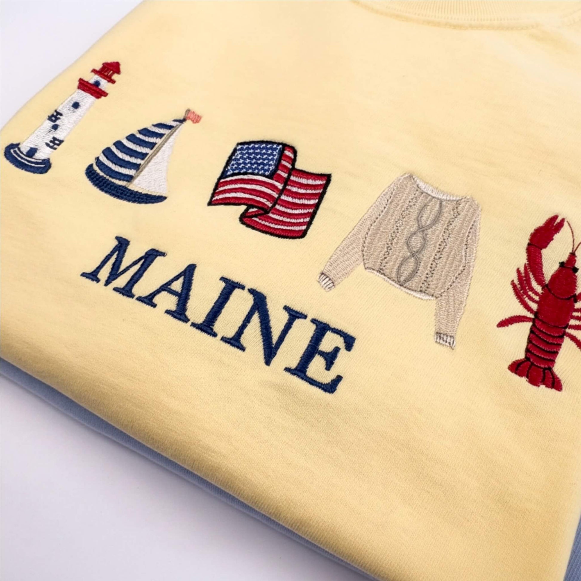A close-up of a Comfort Colors embroidered T-shirt in the color butter featuring nautical-themed embroidery including a lighthouse, sailboat, American flag, sweater, lobster, and the word 'MAINE' in navy lettering.  Part of the Coastal Collection.  Coastal location can be personalized with your favorite location.