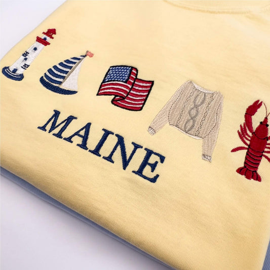 A close-up of a Comfort Colors embroidered T-shirt in the color butter featuring nautical-themed embroidery including a lighthouse, sailboat, American flag, sweater, lobster, and the word 'MAINE' in navy lettering.  Part of the Coastal Collection.  Coastal location can be personalized with your favorite location.