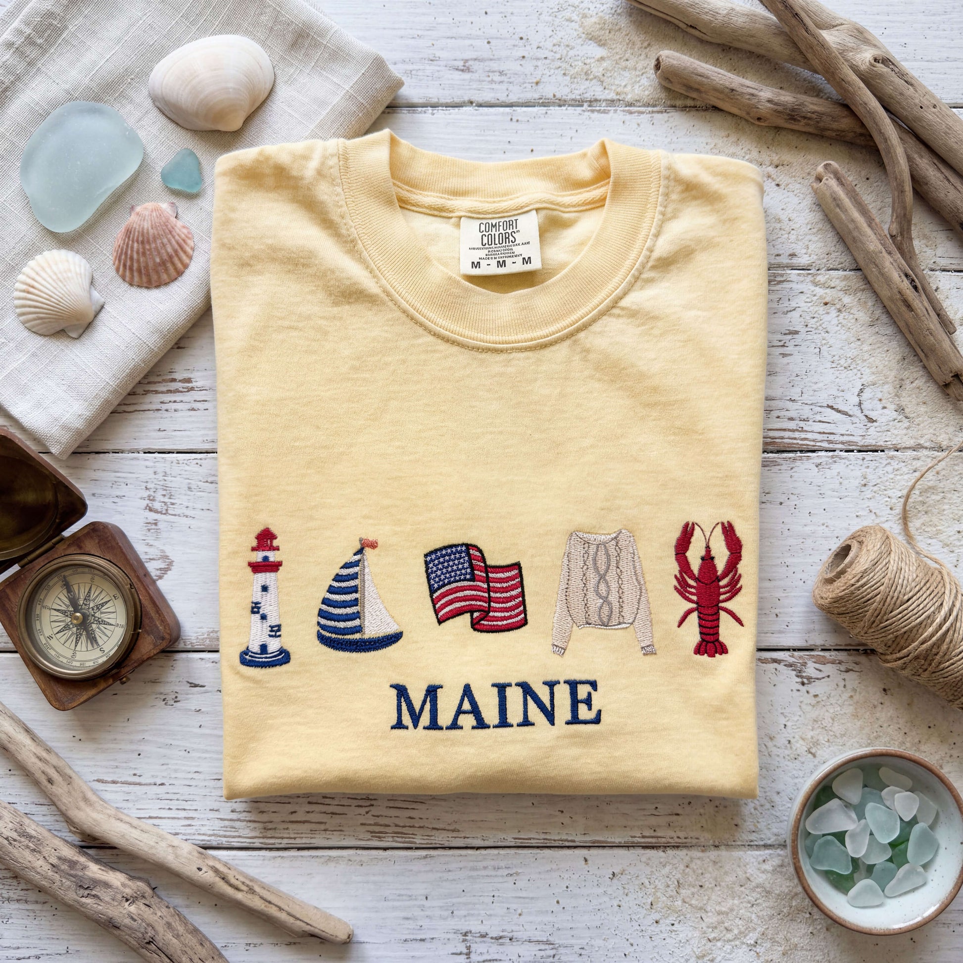 Personalized embroidered Coastal Collection Comfort Colors t-shirt flat lay featuring a coastal-inspired design, styled with beach accessories for a seaside summer look.  Shirt is in the color butter.