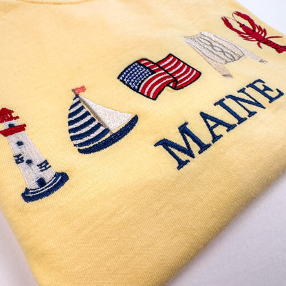A close-up of a Comfort Colors embroidered T-shirt in the color butter featuring nautical-themed embroidery including a lighthouse, sailboat, American flag, sweater, lobster, and the word 'MAINE' in navy lettering.  Part of the Coastal Collection.  Coastal location can be personalized with your favorite location.