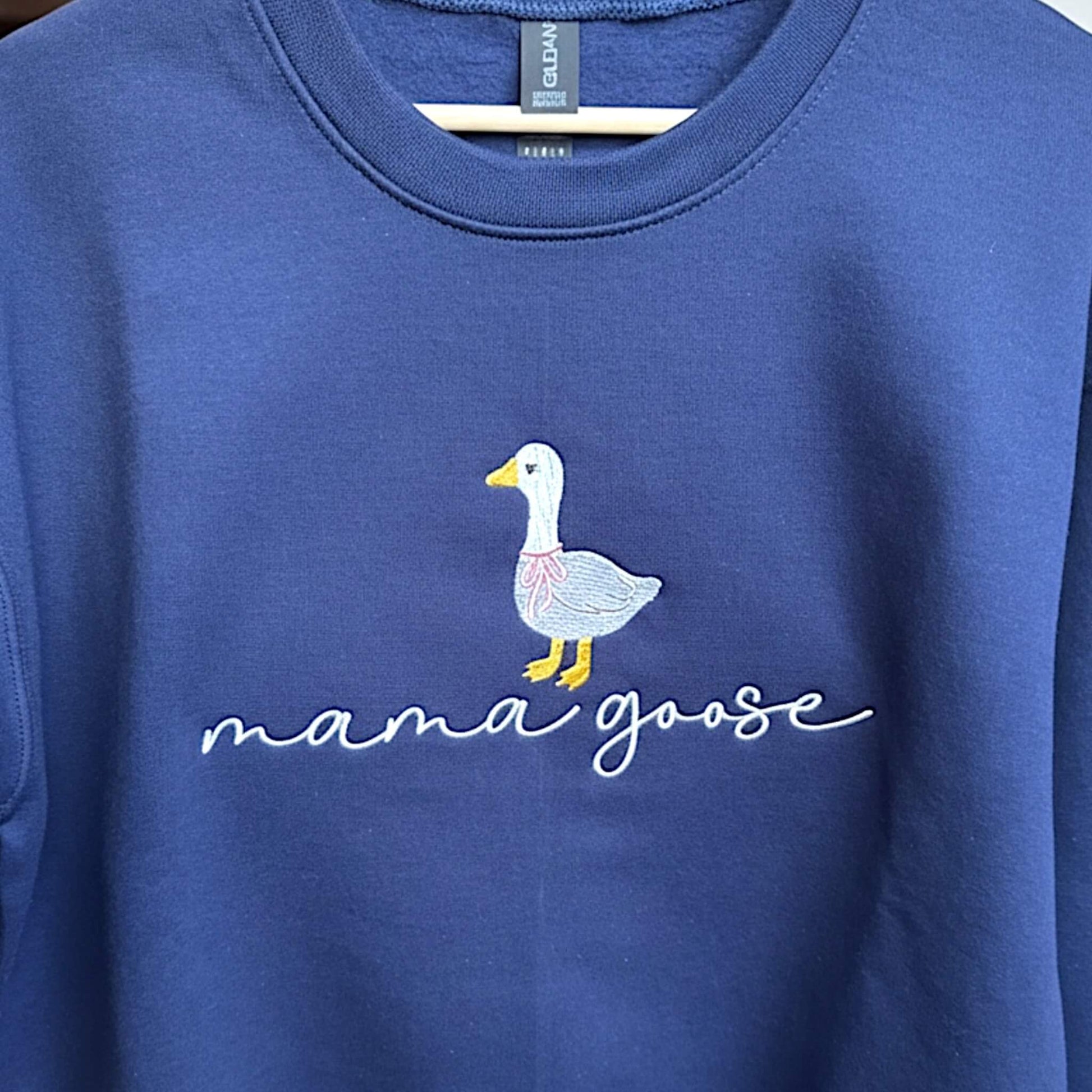 Gildan Softstyle Navy Sweatshirt with an embroiderered design of a cute goose with a pink bow around its neck and the words "mama goose" in a script font.  Picture shows a close up of the embroidery.