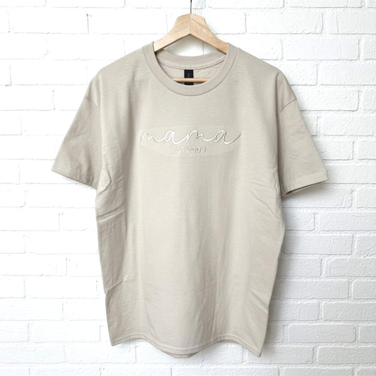 Gildan Softstyle Midweight Sand T-Shirt.  The word mama embroidered in an elegant script font in a similar color of the shirt to create a tone on tone look.  Personalize it with the year you became a mom, by adding Est (your year) underneath the word mama.  Shirt is a neutral color.