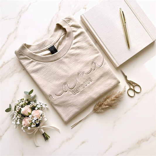 Gildan Softstyle Midweight Sand T-Shirt.  The word mama embroidered in an elegant script font in a similar color of the shirt to create a tone on tone look.  Personalize it with the year you became a mom, by adding Est (your year) underneath the word mama. 