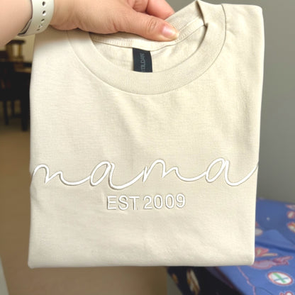 Gildan Softstyle Midweight Sand T-Shirt.  The word mama embroidered in an elegant script font in a similar color of the shirt to create a tone on tone look.  Personalize it with the year you became a mom, by adding Est (your year) underneath the word mama.