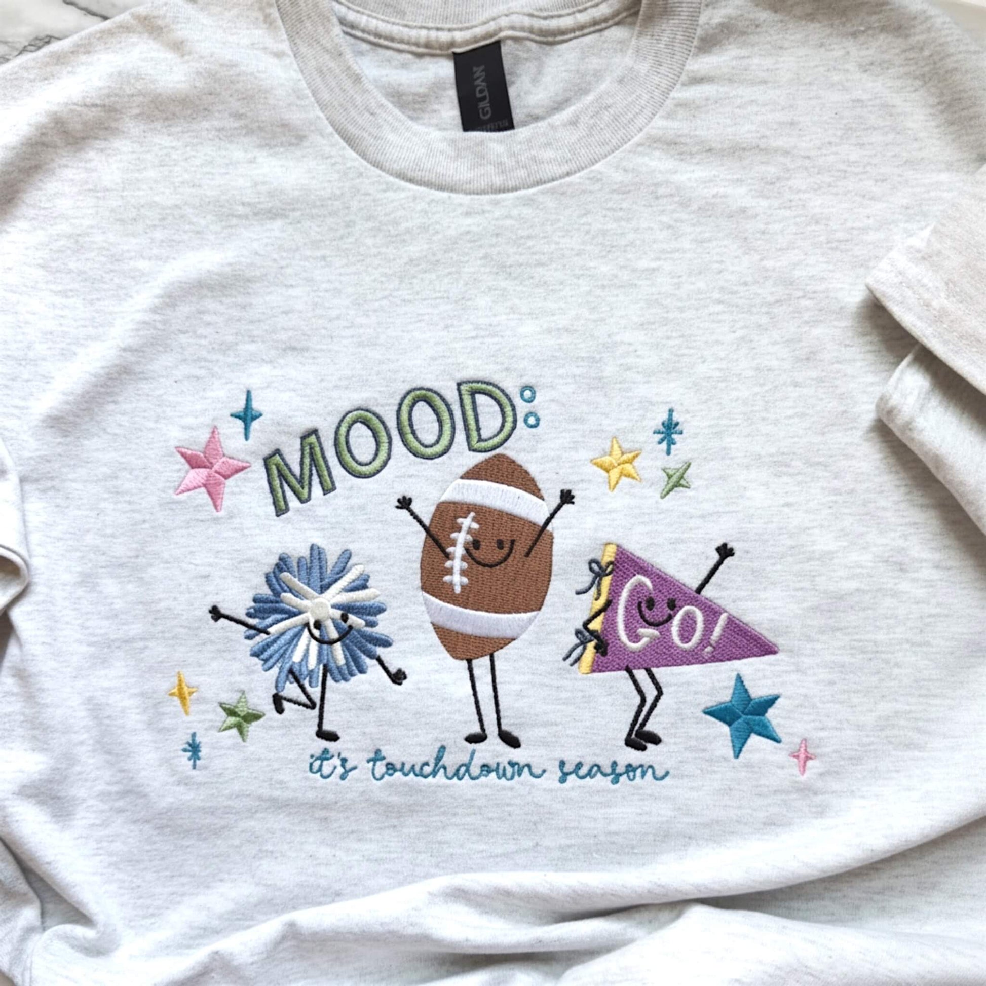 Close-up of colorful embroidered football design featuring pom pom, football and GO banner with stars with the words:  Mood: its football season.  Design is stitched on a Gildan Ash color tee.